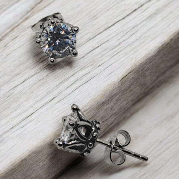 “Close-up of stainless steel crown stud earrings with clear cubic zirconia stones on a light wooden surface.”