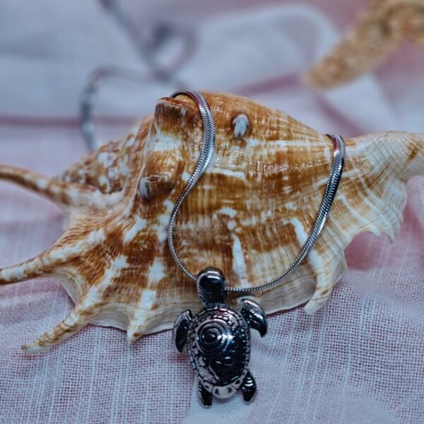 Stainless steel turtle pendant necklace displayed on a seashell with ocean-themed decor on white fabric.