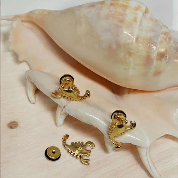 Gold plated stainless steel scorpion stud earrings on white fabric with seashell background