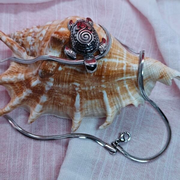 Stainless steel turtle pendant necklace displayed on a seashell with ocean-themed decor on white fabric.